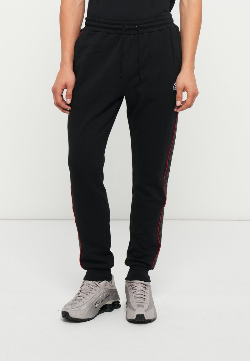 Zwarte joggingsbroeken van zachte stof met rode zijstrepen, voorzien van een elastische tailleband en een trekkoord, gecombineerd met grijze sneakers.
