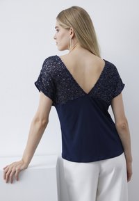 Top blu navy con maniche corte a volants, scollatura decorata di paillettes e design con scollo a V sul retro. Il tessuto del corpo è liscio e si abbina alla parte superiore impreziosita.