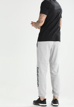 Mężczyzna w szarych spodniach dresowych Adidas, czarnych butach sportowych i czarnej koszulce z krótkim rękawem stojący ze złączonymi rękami za plecami.