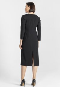 Czarna sukienka do kolan o prostym kroju, z długimi rękawami, rozcięciem z tyłu i subtelnym okrągłym dekoltem. Noszona z czarnymi szpilkami.