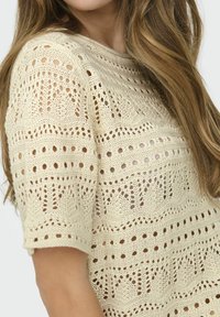 Suéter de punto color crema claro con mangas cortas, que presenta un patrón texturizado de puntos abiertos y agujeros decorativos en todo su diseño.