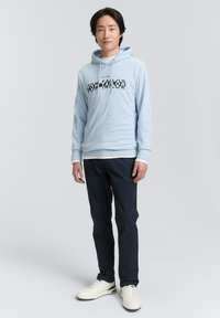 Világoskék kapucnis pulóver fekete grafikai szöveggel, sötét navy nadrággal és fehér sportcipővel párosítva, egy semleges háttér előtt áll.