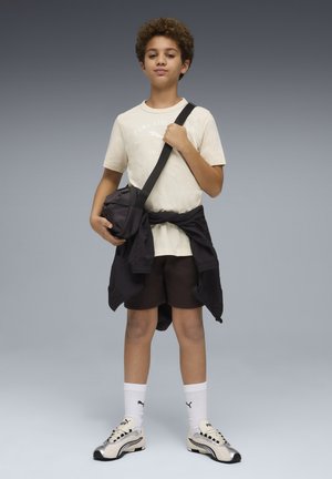 Jasnobeżowa koszulka z nadrukiem, czarne szorty oraz czarna kurtka związana wokół talii. Białe skarpetki i srebrne buty sportowe z czarnymi detalami.