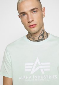Piparmētru zaļš T-krekls ar īsām piedurknēm, kurā ir liels balts "Alpha Industries" logotips un ģeometrisks dizains. Mīksta auduma tekstūra.