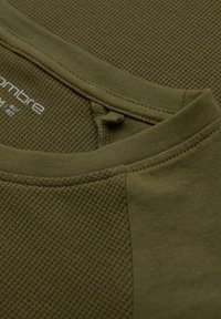 Oliivivihreä paita, jossa on teksturoitu kuvio etuosassa, sileä kangas hihoissa ja pyöreä pääntie. Sisältää pienen tarradetailin.