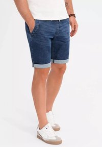 Shorts de mezclilla en azul oscuro, con dobladillo enrollado, bolsillos frontales y una textura suave. Combinados con zapatillas grises y blancas.