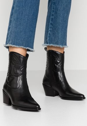 Czarne kowbojki z skóry licowej do kostki, z haftowanymi wzorami, obcasami typu słupek i zamkami po bokach, noszone z niebieskimi, przyciętymi dżinsami z postrzępionym dołem.