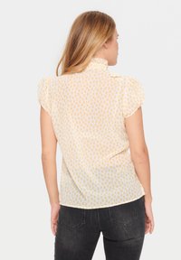 Korte mouwen blouse van doorschijnende gele stof met lichtbruine luipaardprint, met een hoge kraag en geplooide accenten op de schouders.