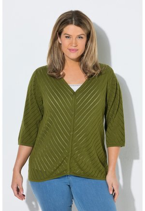 Vrouw met schouderlang haar draagt een olijfgroene gebuttonede cardigan met diagonale strepen en lichtblauwe spijkerbroek, staand tegen een witte muur.
