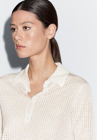 Witte, semi-doorzichtige gebreide polo met kraag, voorzien van een textuurpatroon en knoopsluiting. Zacht materiaal met een ontspannen pasvorm.