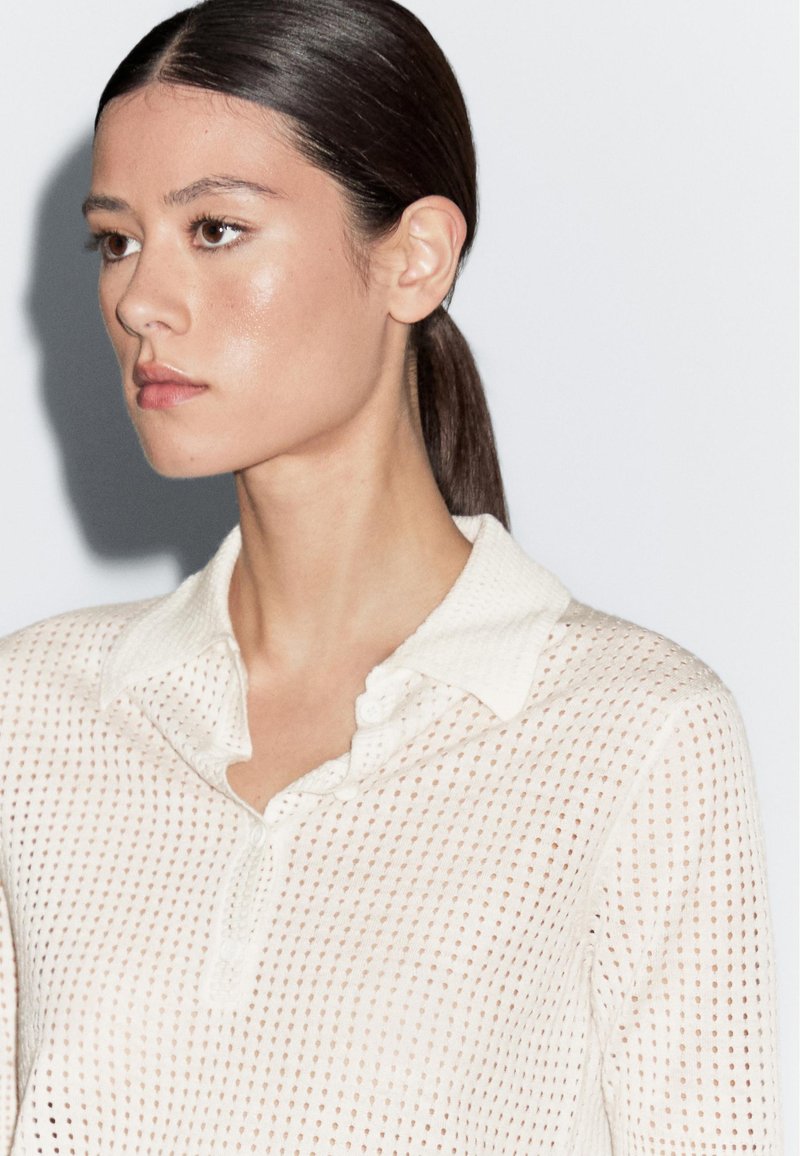 Witte, semi-doorzichtige gebreide polo met kraag, voorzien van een textuurpatroon en knoopsluiting. Zacht materiaal met een ontspannen pasvorm.