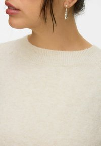 Skræddersyet cremefarvet strikket sweater med en tekstureret overflade og ribbet rund halsudskæring. Sølvfarvede hængende øreringe med sfæriske accenter er delvist synlige.