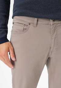 Pantaloni beige con una superficie liscia, dotati di una tasca frontale e accessori metallici. Indossati con una camicia a maniche lunghe a fantasia blu scuro.