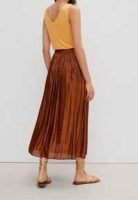 Top arancione senza maniche con scollatura a V profonda, abbinato a una lunga gonna plissettata e lucida di colore marrone e sandali beige con cinturini.