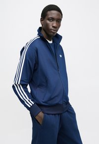 Tmavomodrá tréningová bunda z hladkej látky s tromi bielymi pruhmi na rukávoch, so zipsovým uzáverom a prednými vreckami. Obsahuje malé logo.