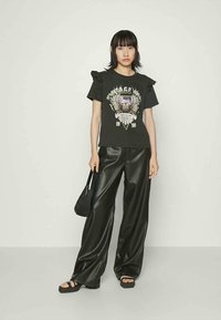 Zwarte t-shirt met grafisch ontwerp met vleugels en tekst, gecombineerd met losse zwarte faux leren broek en zwarte sandalen. Accessoires: kleine tas.