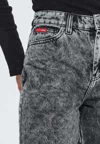 Gros plan sur un jean en denim noir délavé avec une étiquette rouge sur la petite poche avant, porté avec un haut noir à manches longues.