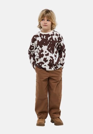 Enfant blond debout, les mains dans les poches, portant un pull imprimé tache de vache marron et blanc, un pantalon cargo marron et des chaussures beige sur fond blanc.