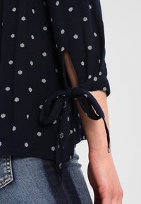 Blusa blu navy con motivo a diamante bianco, tessuto testurizzato e maniche annodate che rivelano un accenno di pelle. Indossata sopra dei jeans.