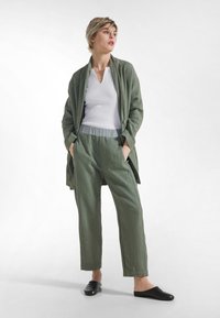 Lekki, teksturowany oliwkowo-zielony strój z długą kurtką i luźnymi spodniami, zestawiony z białym topem i czarnymi wsuwanymi butami.