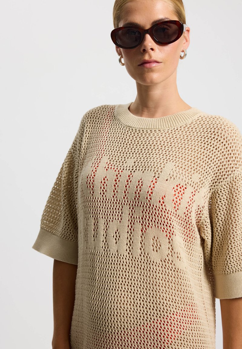 Pull en maille beige avec une coupe ample, manches courtes, et un graphique rouge audacieux "thinking" sur le devant. Encolure et ourlet lisses.