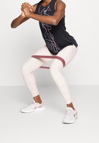 Top negro sin mangas con logo blanco y rosa, leggings texturizados en beige claro y zapatillas blancas con acentos rosas. Banda de resistencia posicionada en los muslos.