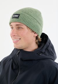 En mössa i ljusgrön ribbad stickning med en svart SLOPE-logopus, bärs tillsammans med en svart zip-up-jacka med huva och ventilationsdetaljer.