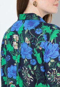 Donna vista di lato che indossa una camicia blu navy con grandi motivi floreali blu e bianchi e orecchini a cerchio dorati.