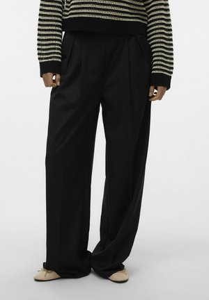 Pantalons noirs à jambes larges avec des plis, fabriqués dans un tissu lisse. Dotés d'une taille haute et d'une coupe décontractée, associés à des chaussures plates de couleur claire.
