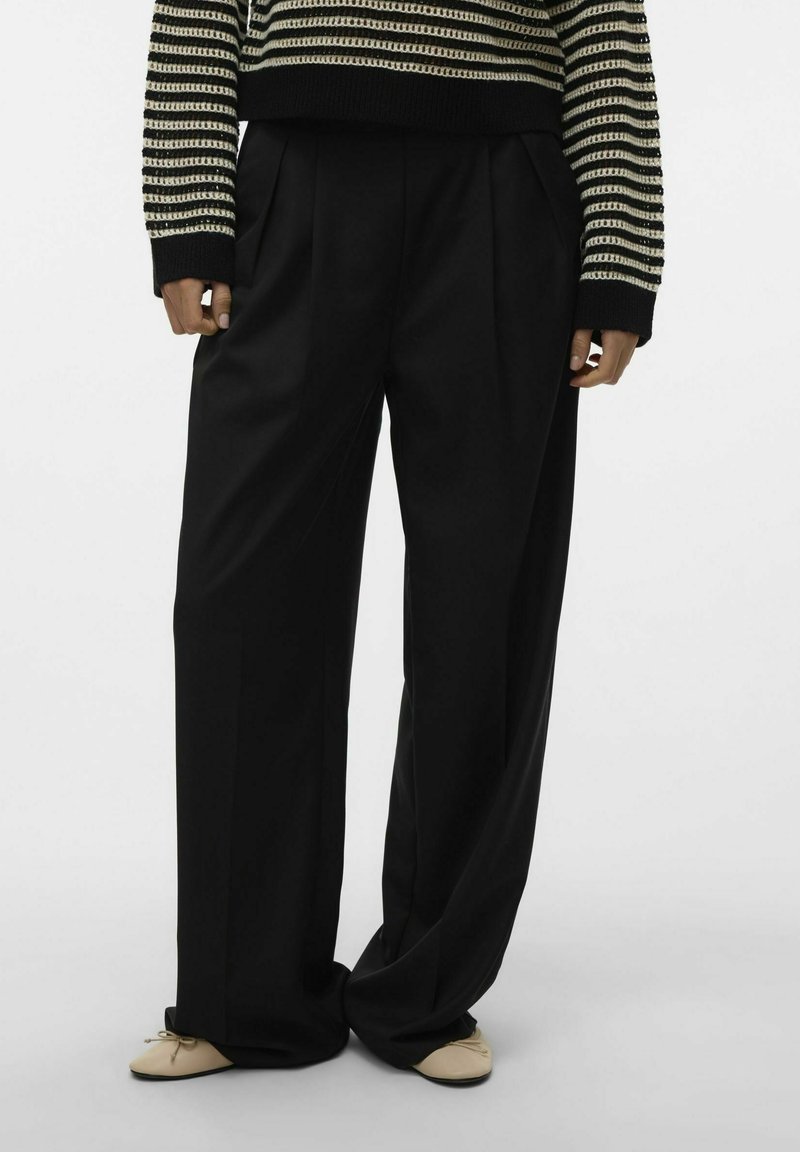Pantalons noirs à jambes larges avec des plis, fabriqués dans un tissu lisse. Dotés d'une taille haute et d'une coupe décontractée, associés à des chaussures plates de couleur claire.