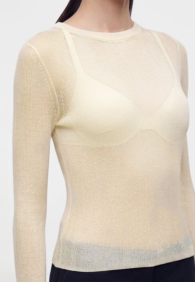 Femme portant un haut en maille beige transparent à manches longues laissant apparaître un soutien-gorge beige assorti en dessous, sur un fond uni.