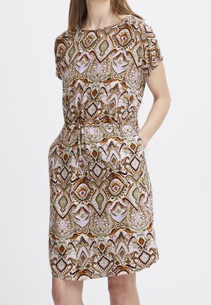 Kobieta w sukience o krótkich rękawach sięgającej do kolan, z misternym geometrycznym i kwiatowym wzorem w odcieniach brązu, beżu i różu, z rękami w kieszeniach.