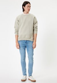 Felpa grigia chiara con maniche raglan, polsini a coste e scollo rotondo, abbinata a jeans azzurri e sneakers bianche con dettagli marroni.