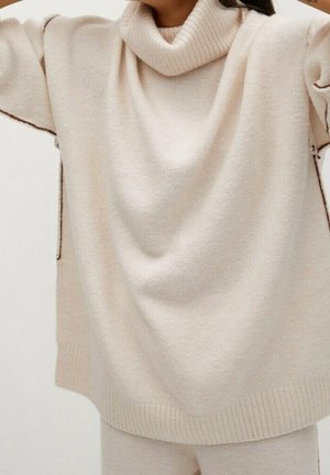 Femme portant un pull col roulé en tricot crème surdimensionné avec des détails de coutures visibles le long des manches et du corps, associé à un pantalon assorti.