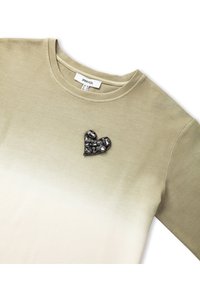 Chemise à manches longues en olive clair avec un dégradé vers le beige. Dispose d'un patch en forme de cœur noir orné sur la poitrine. Matériau en mélange de coton.