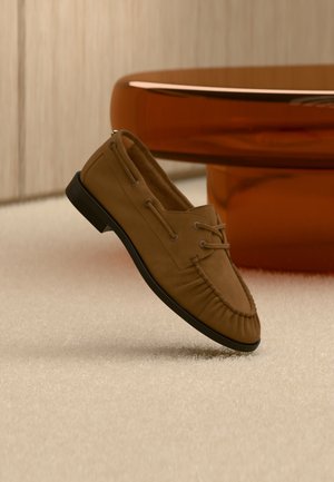 Mocassin en cuir suédé marron avec semelle noire flottant au-dessus d'un tapis beige, avec une table ronde orange floutée en arrière-plan.
