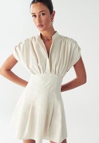 Kratký rukáv, svetlo béžové šaty s vysokým golierom, plisovanou sukňou a nariaseným detailom tkaniny v oblasti pása. Hladká textúra, minimalistický dizajn.