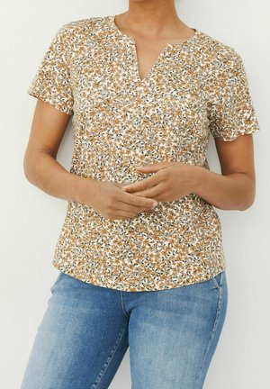 Kurzärmeliges Floral-Blusenshirt in Beige, Weiß und Schwarz. Mit einem gespaltenen Halsdesign und einem strukturierten Stoff. Kombiniert mit blauen Denim-Jeans.