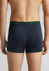 Man som bär mörkblå boxerkalsonger med en mörkgrön midja, sedd bakifrån, ståendes mot en neutral bakgrund.