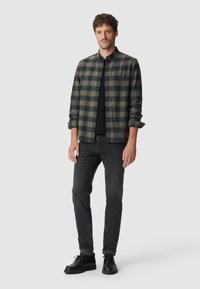 Man in een open groene en zwarte geruite overhemd over een zwart T-shirt, donkere grijze jeans en zwarte leren schoenen, staand tegen een witte achtergrond.