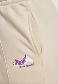 Beige Sweatpants mit einer Seitentasche und einem lila Berggrafik mit dem Text "JUST RHYSE" darunter. Weicher Stoff.
