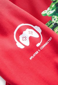 Tissu rouge présentant un graphique blanc d'une manette de jeu avec des écouteurs et le texte "#JOUEUR 1 - CONNECTÉ" en dessous.