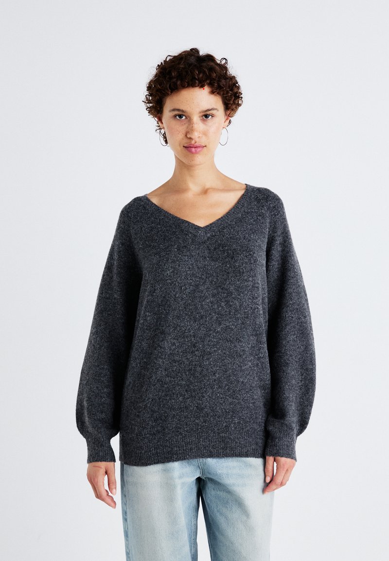Maglione grigio scuro a scollo a V con maniche lunghe e a palloncino, realizzato in un materiale morbido e testurizzato, abbinato a jeans azzurri chiari.