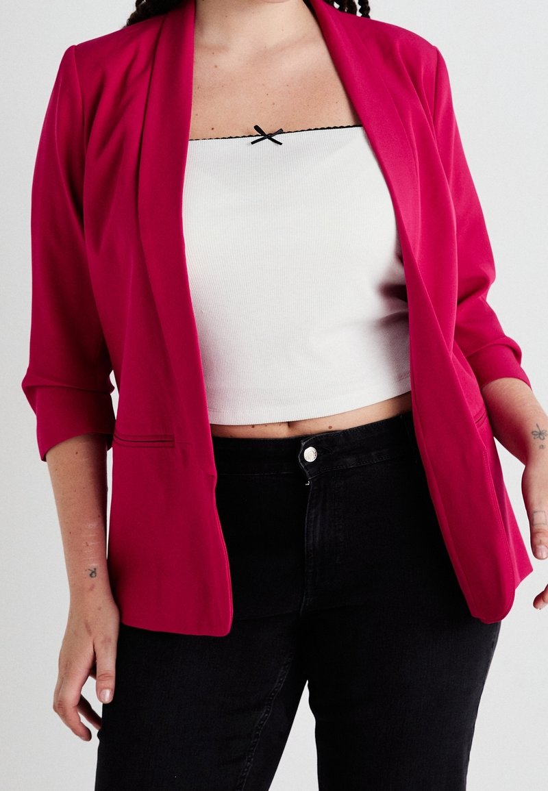 Fuchsia blazer med trekvartsärmar, öppen front och två framfickor, lagd över en vit kort topp och svarta högmidjade jeans.