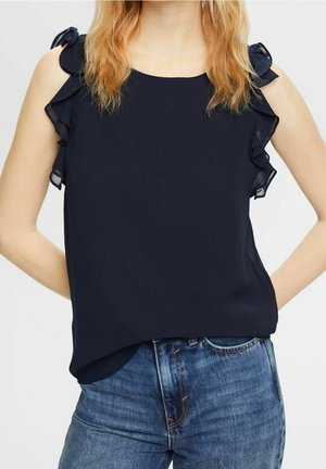 Mujer con cabello castaño claro lleva una blusa negra sin mangas con lazos fruncidos en los hombros y vaqueros azules de mezclilla contra un fondo liso.