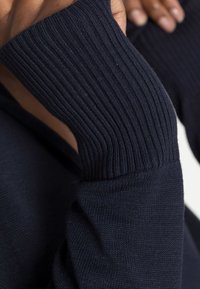 Pull bleu marine avec poignets côtelés et texture lisse du corps, présentant des coutures insérées et une coupe ajustée.