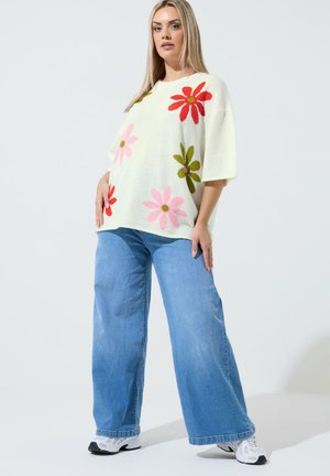 Woman wearing white oversized sweater with large pink, red, and green flower patterns, wide-leg blue jeans, and white sneakers.