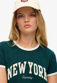Camiseta verde con cuello crema, que presenta "NUEVA YORK" en texto blanco en negrita y un logotipo de "Superdry". Acompañada de una gorra crema con un parche.