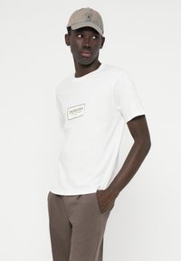 Biely tričko s čiernym obdĺžnikovým logom, skombinované s hnedými nohavicami a béžovou čiapkou so malým emblémom. Jednoduchý, neformálny dizajn.