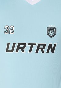 Ανοιχτό μπλε αθλητικό μπλουζάκι με λευκό V-ντεκολτέ. Περιλαμβάνει μαύρους αριθμούς "32" και λογότυπο με αρκούδα και τρία αστέρια, προσανατολισμένο μπροστά. Το κείμενο αναγράφει "URTRN."